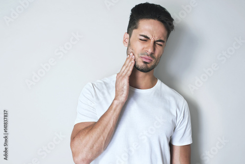 Portrait young man touch cheek. He has strong toothache and suffering from pain wearing casual white t-shirt against grey background. Feel toothache