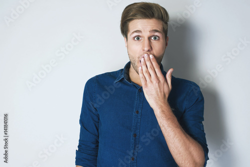 Young attractive man in blue shirt amazed in shock covering his mouth with hand and looking at camera against grey background. The sign of silence