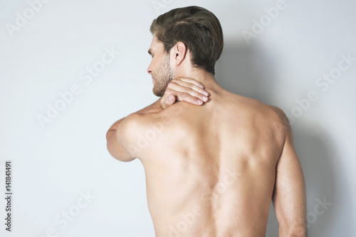 View on man's nude back, close up. Back view portrait of a muscular young man with neck pain against grey background