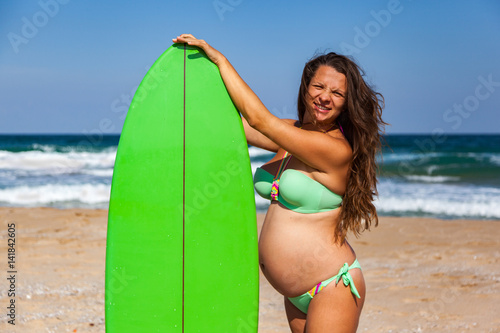 Young pretty pregnant woman is standing on the beach of Black sea with her green surfboard