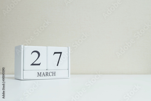 Closeup white wooden calendar with black 27 march word on blurred white wood desk and cream color wallpaper in room textured background with copy space , selective focus at the calendar