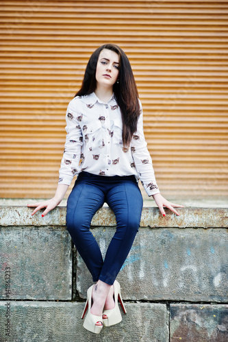Young stylish brunette girl on shirt, pants and high heels shoes, sitting and posed background orange shutter. Street fashion model concept.
