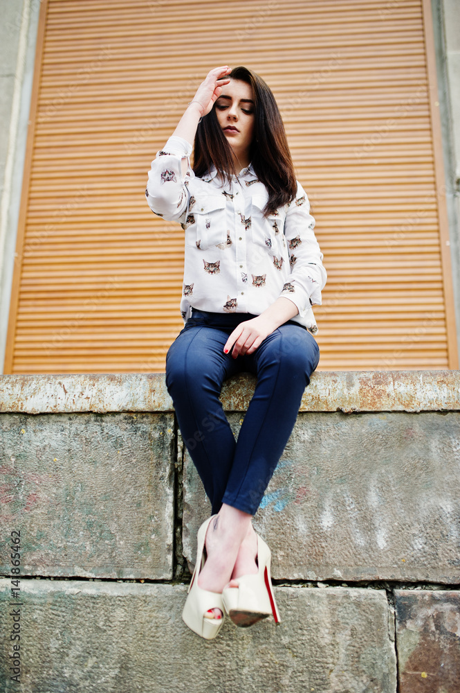 Young stylish brunette girl on shirt, pants and high heels shoes, sitting and posed background orange shutter. Street fashion model concept. Young stylish brunette girl on shirt, pants and high heels shoes, sitting and posed background orange shutter. Street fashion model concept.