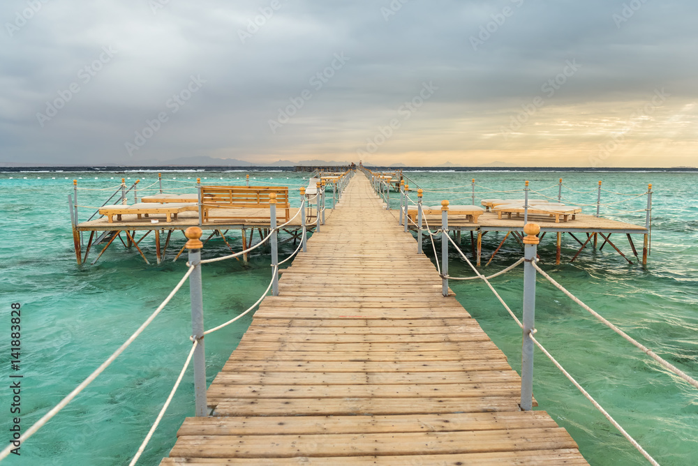 Obraz premium Pier with chaise longues in the sea in resort at sunset. Summer vacation. View at a clear sea with turquoise water. Summer vacation at a sea coastline in a exotic country.