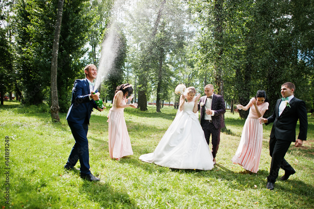Fashionable wedding couple with bridesmaids and best mans with explosion of champagne bottle at forest on sunny wedding day.