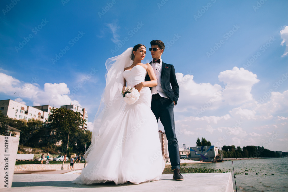 Elegant smiling young bride and groom walking on the beach, kissing and having fun, wedding ceremony near the lake