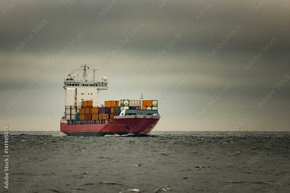 Red cargo container ship sailing from Baltic sea in cloudy day
