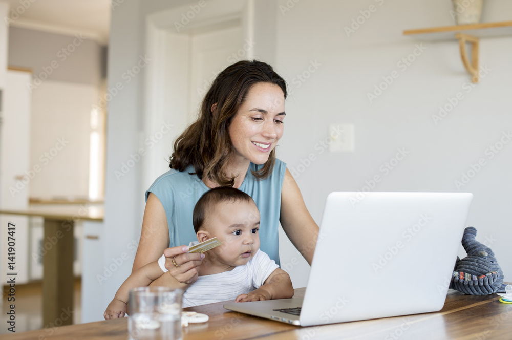 Woman with baby boy online shopping while using laptop computer at home Woman with baby boy online shopping while using laptop computer at home