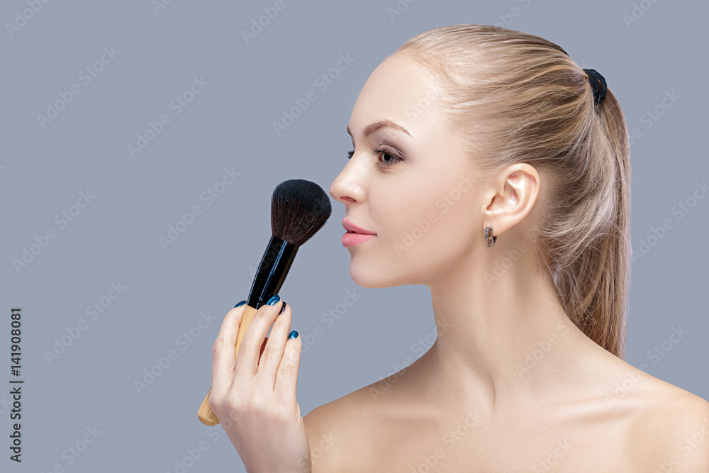 beautiful blond woman holding makeup brush on a gray background.