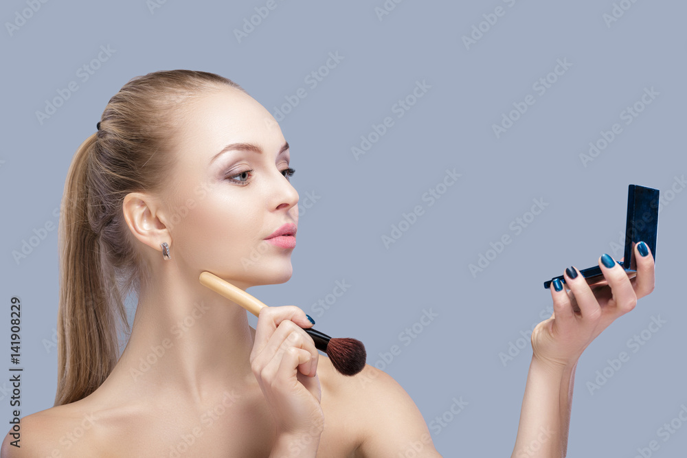 Obraz premium beautiful blond woman holding makeup brush and looking in the mirror on a gray background. 