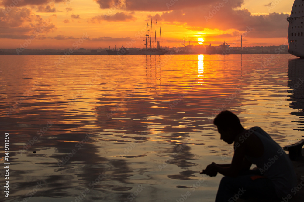 Silhouette of man fishing by hand and sail boat at sunrise in Havana port, Cuba