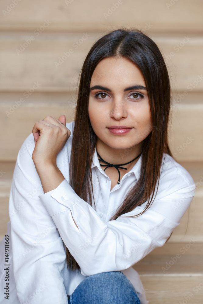 Closeup portrait of smiling Caucasian brunette young beautiful girl woman model with long dark hair and brown eyes in white office shirt sitting on staircase looking in camera Closeup portrait of smiling Caucasian brunette young beautiful girl woman model with long dark hair and brown eyes in white office shirt sitting on staircase looking in camera