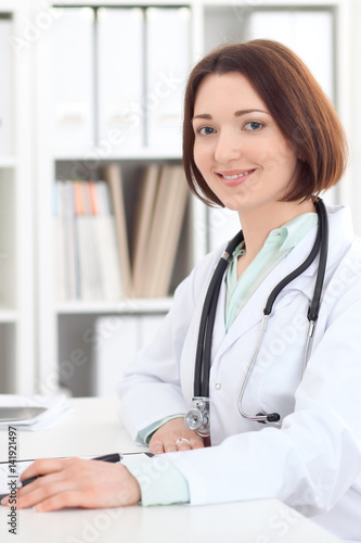 Young brunette female doctor sitting at the table and working at hospital office. Health care, insurance and help concept. Physician ready to examine patient