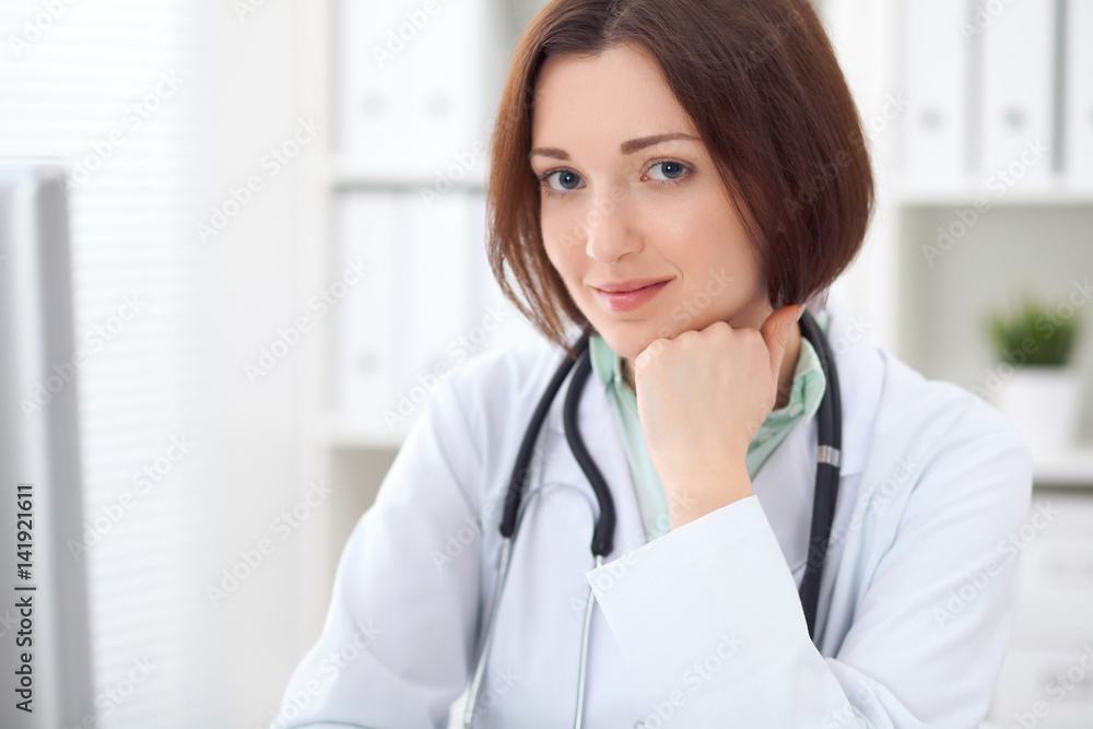 Young brunette female doctor sitting at a desk and working on the computer at the hospital office. Health care, insurance and help concept. Physician ready to examine patient Young brunette female doctor sitting at a desk and working on the computer at the hospital office. Health care, insurance and help concept. Physician ready to examine patient