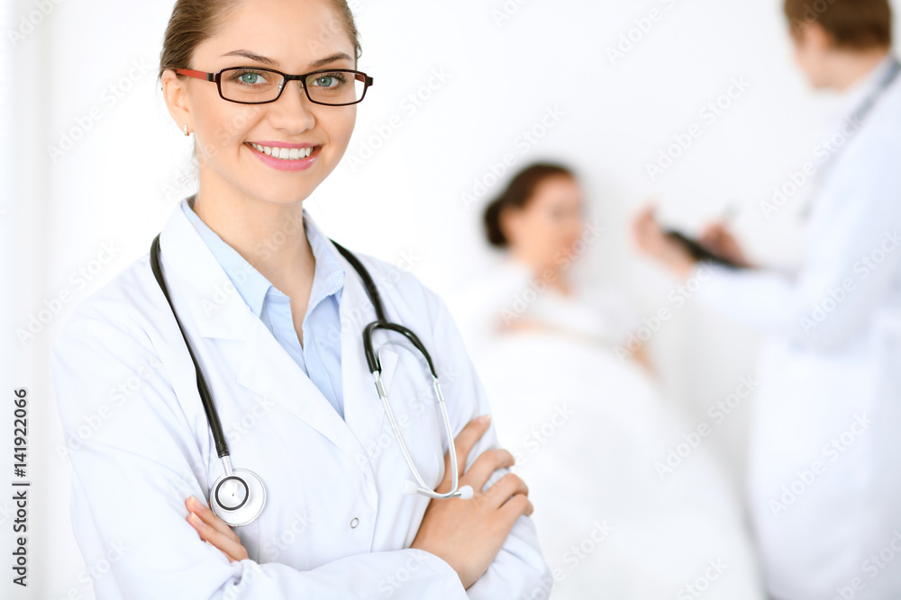 Cheerful smiling female doctor on the background with physician and his patient in the bed. High level and quality of medical service concept. Cheerful smiling female doctor on the background with physician and his patient in the bed. High level and quality of medical service concept.
