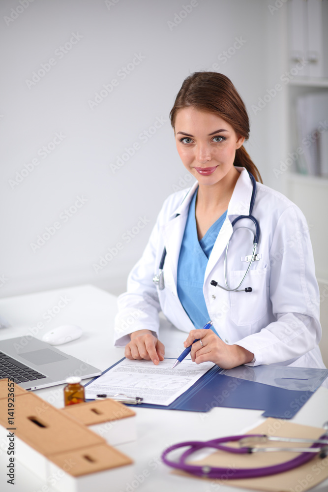 Beautiful young smiling female doctor sitting at the desk and writing Beautiful young smiling female doctor sitting at the desk and writing