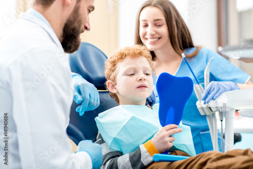 Młoda chłopiec patrzeje lustro z toothy uśmiechu obsiadaniem na krześle z dentystą i asystentem przy stomatologicznym biurem