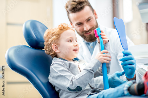 Young excited boy sitting on the chair holding big toothbrush with doctor in the dental office