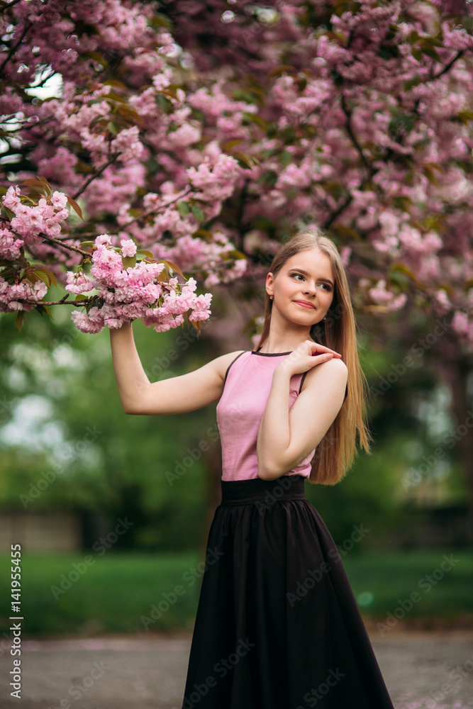 Charming girl in pink blouse and black skirt poses for photographers on the background of beautiful flowering trees. Spring. Sakura. Charming girl in pink blouse and black skirt poses for photographers on the background of beautiful flowering trees. Spring. Sakura.