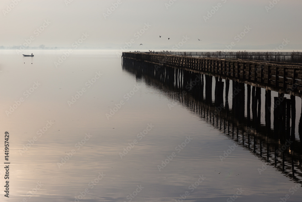 A pier perfectly reflecting on water, with some birds fying, and little fisherman boat at some distance on the opposite side.