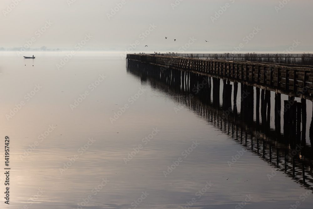 Fototapeta premium A pier perfectly reflecting on water, with some birds fying, and little fisherman boat at some distance on the opposite side.
