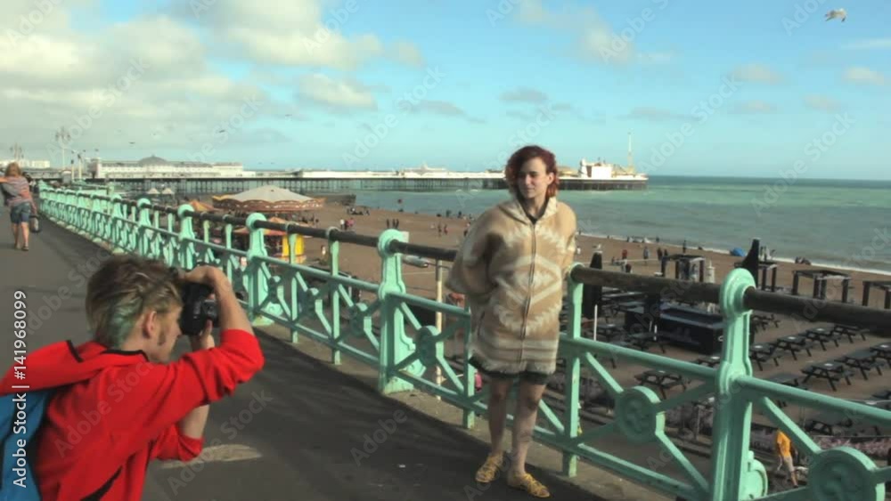 UK, Brighton. UK, Brighton. Young happy couple taking photo on the coast. Happy lovers, woman and man traveling on vacation on ocean. Girl leaning on railing of beach promenade. UK, Brighton. UK, Brighton. Young happy couple taking photo on the coast. Happy lovers, woman and man traveling on vacation on ocean. Girl leaning on railing of beach promenade.