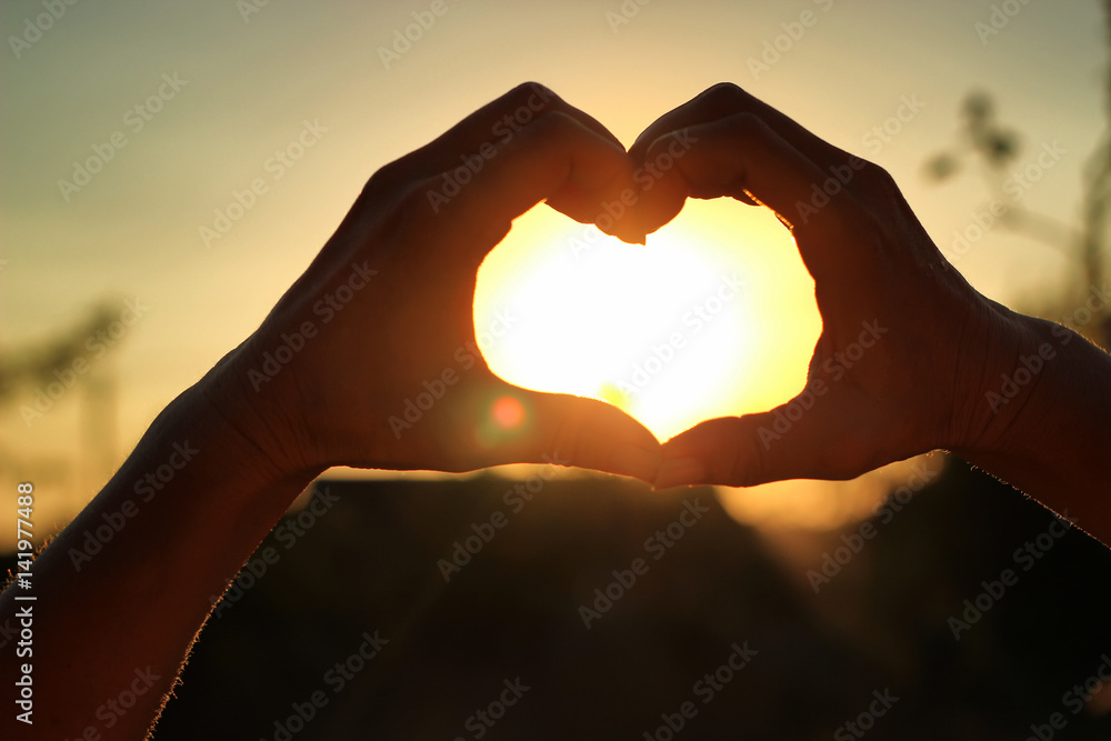 Fototapeta premium Women's hands are crossed in the form of the heart through which the sun' rays make the way at the sunset