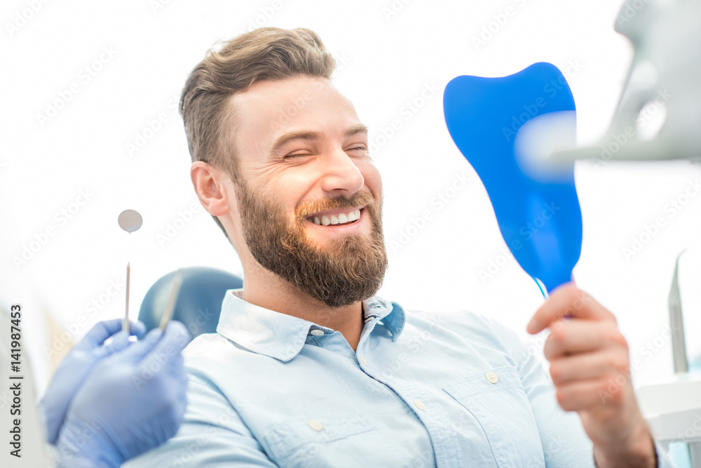 Handsome male patient looking at his beautiful smile sitting at the dental office - obrazy, fototapety, plakaty 