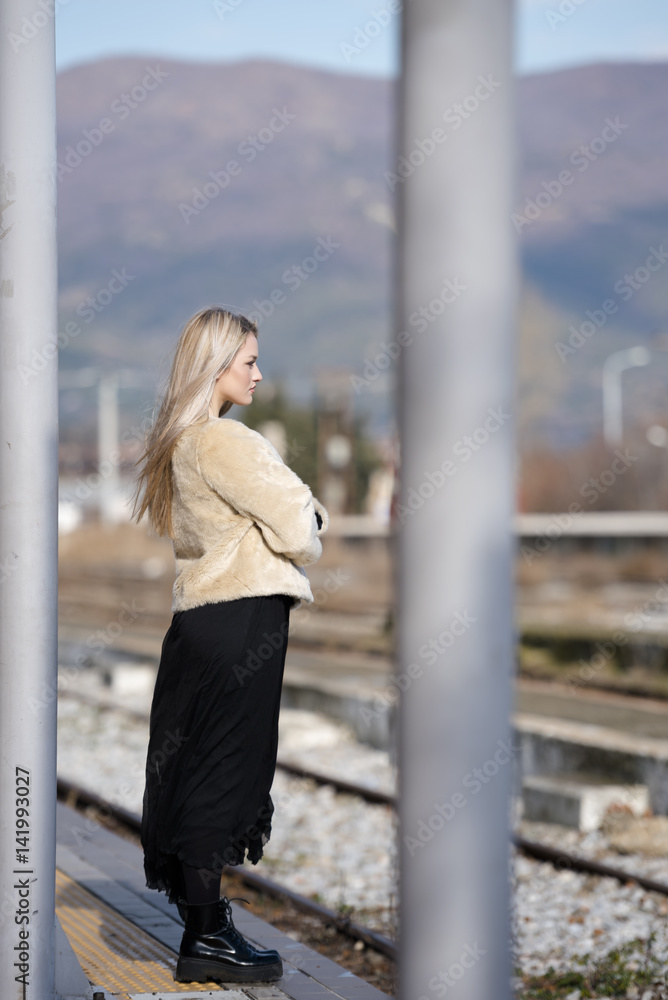 Blond young woman standing at the edge railway platform waiting wear sheep jacket and black long skirt