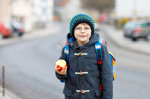 Little kid boy with eye glasses walking from the school and eating apple photo