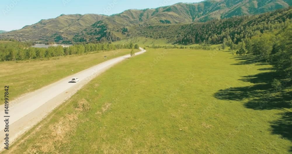 Aerial UHD 4K view. Drone flight over cars on a winding road in the hills and meadow at sunny summer morning. Green trees, fresh river and sun rays on horizon. Rural highway below. Aerial UHD 4K view. Drone flight over cars on a winding road in the hills and meadow at sunny summer morning. Green trees, fresh river and sun rays on horizon. Rural highway below.