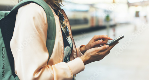 Enjoying travel. Young woman waiting on station platform with backpack on background electric train using smartphone. Tourist texting message and plan route of railway, railroad transport concept