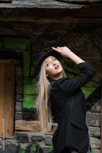 Cute blond teen with long straight hair wear black jacket and hat standing against stone wall with big window