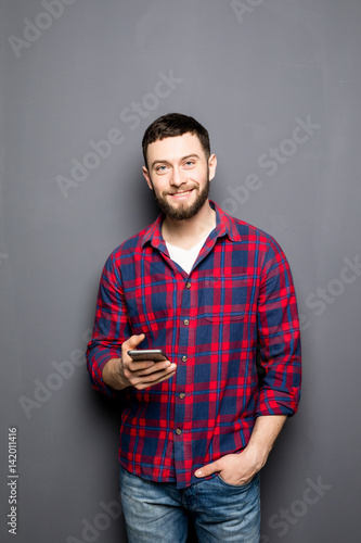 Confident young man holding smart phone and looking at it while standing against grey background