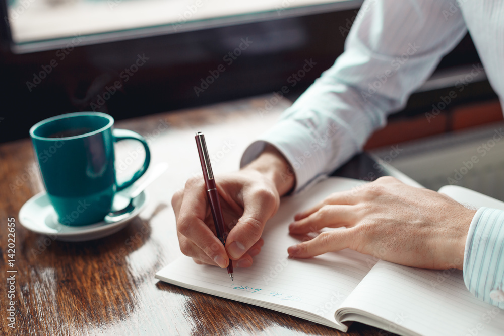 Close up of hands of a businessman writing in a notebook and a cup of coffee. A freelance man is working in a cafe. Coffee break. Close up of hands of a businessman writing in a notebook and a cup of coffee. A freelance man is working in a cafe. Coffee break.