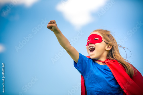 Funny little girl playing power super hero over blue sky background. Superhero concept.