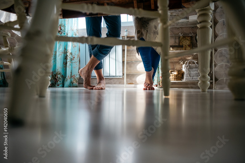 Cropped image of man and woman standing near a large wooden table in the kitchen. Human legs wearing jeans and table and chair legs. Place for text