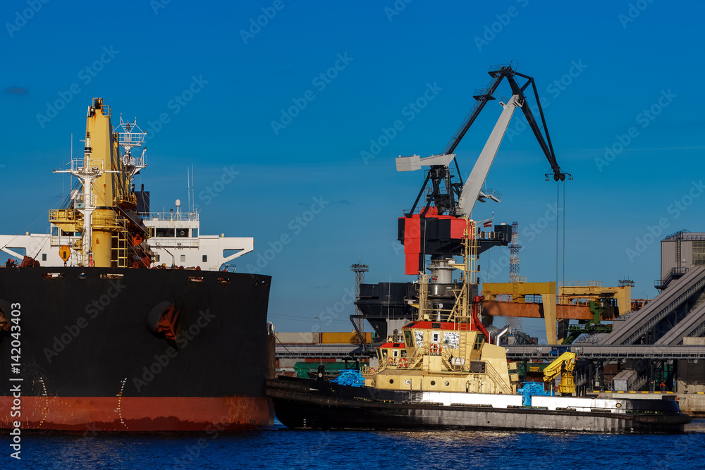Fototapeta premium Black cargo ship mooring at the port with tug ship support