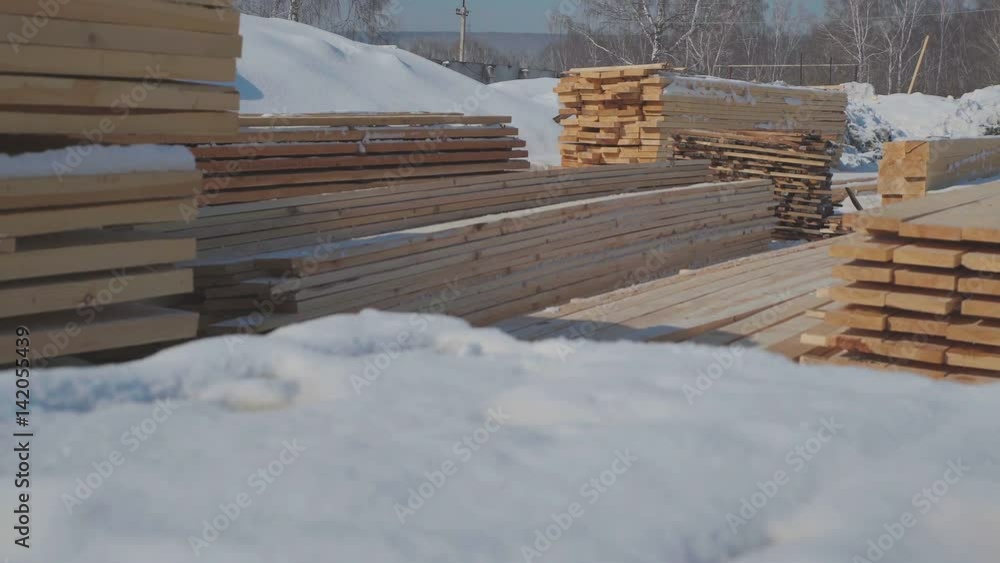 Wooden blanks for the production of glued timber stand in the street in neat rows. Wooden beams and boards neatly stacked. Boards for building standing on the street on a background of white snow. Wooden blanks for the production of glued timber stand in the street in neat rows. Wooden beams and boards neatly stacked. Boards for building standing on the street on a background of white snow.