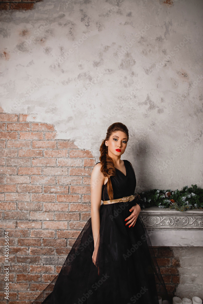 beautiful brunette girl in long black dress makeup red lips and manicure is standing near the fireplace looking down