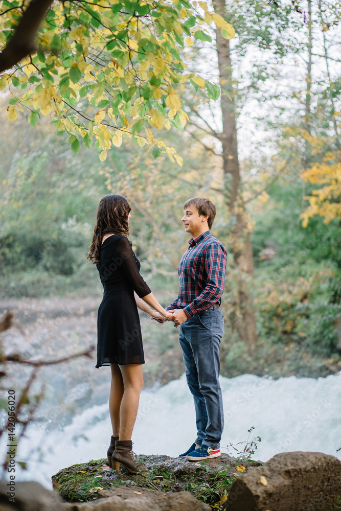 pair of beautiful brunette with a guy holding hands in nature at the waterfall fall trees