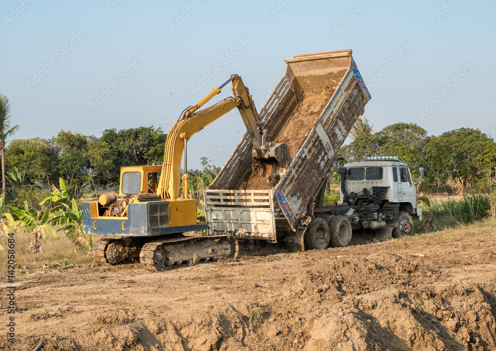 Yellow backhoe digging soil fill in rear truck Yellow backhoe digging soil fill in rear truck
