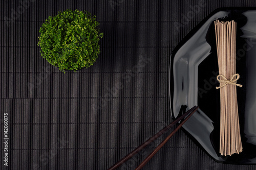 Bundle raw asian noodles in plate with chopsticks, green plant on black striped mat background with copy space, top view. Modern minimalistick style restaurant menu mock up. photo