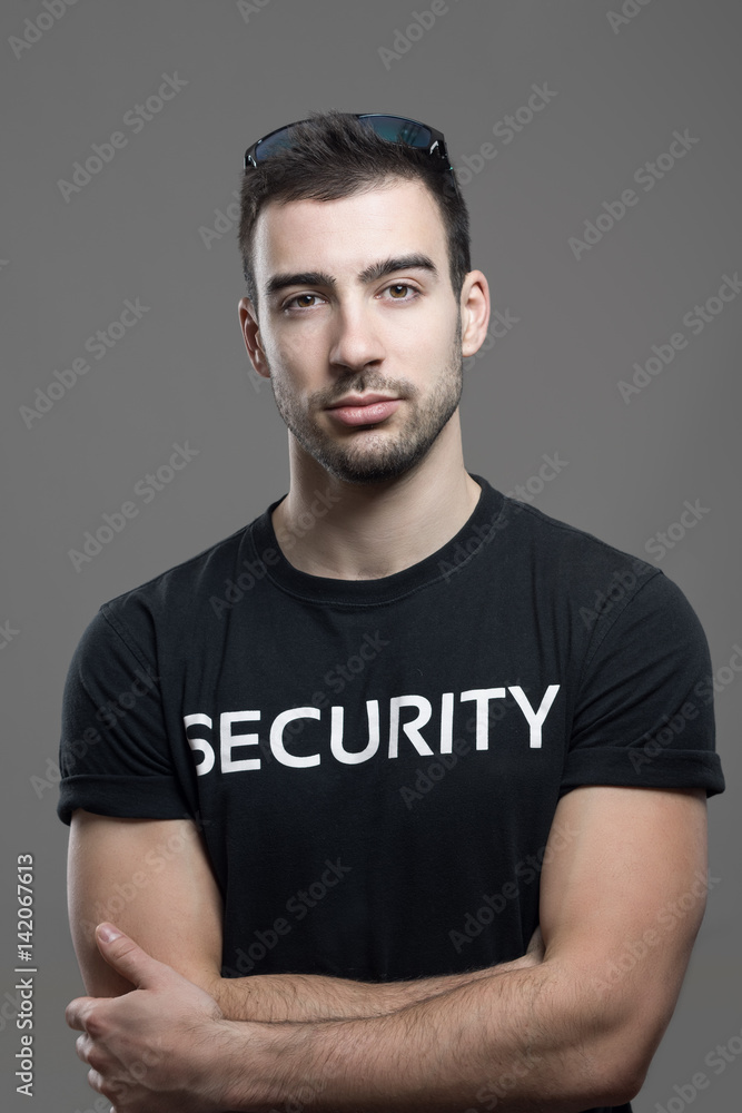 Cool fit security guard in black shirt with crossed arms looking at camera. Atmospheric contrasty portrait over gray studio background.