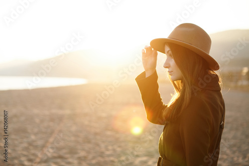 Profile of charming young female in hat and coat spending nice warm evening outdoors on city beach, spring sun shining brightly in background. Cute girl having rest at seaside adjusting headwear