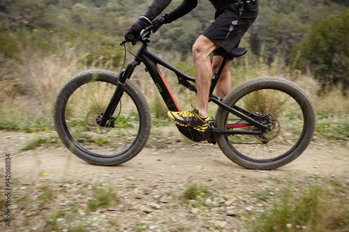 Cropped shot of male cyclist riding black two-wheeled motor-power bicycle with pedal-assist system while cycling on mountain track in rural area. Extreme, sports, fitness and healthy lifestyle