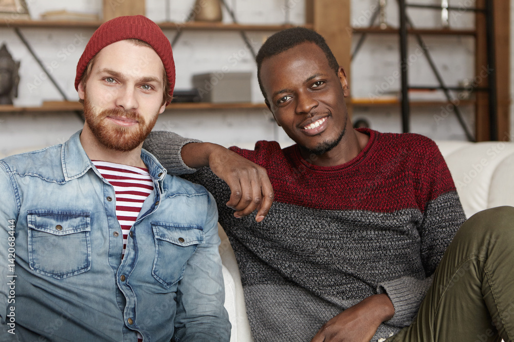 Interracial friendship concept. Happy African American male in casual  sweater resting elbow on his best friend's shoulder while sitting on white  couch at coffee shop, talking and having fun together Stock Photo |