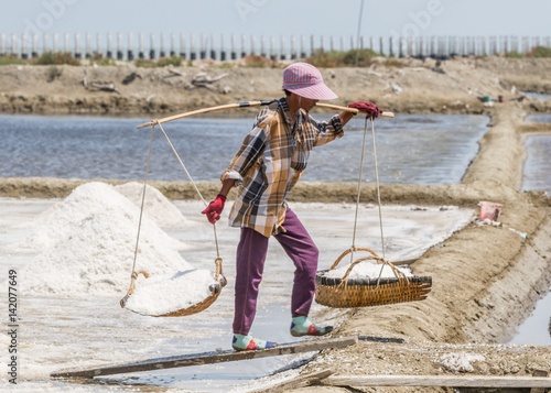 Salt field worker carrying salt with traditional shoulder pole with baskets during salt harvest in Ban Laem, Thailand photo