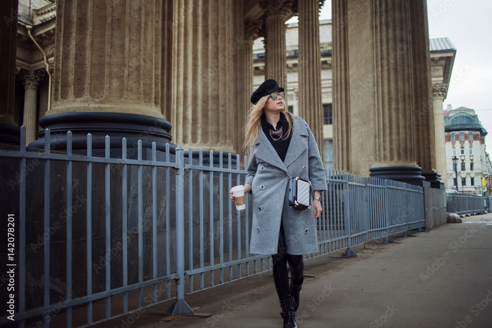 Young, hip and attractive blonde walking around the city with coffee to go, girl in a stylish hat and a gray coat
