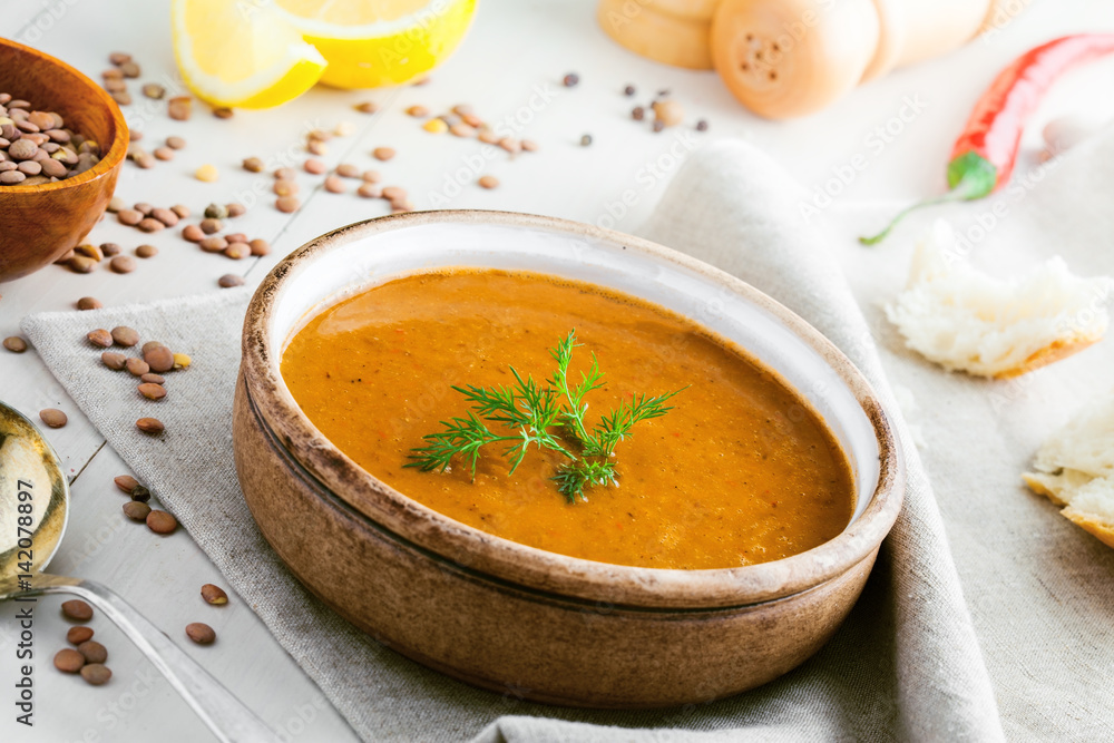 Lentil soup in a rustic bowl with piece of bread on a cotton canvas tablecloth. Healthy vegetarian food on a white wooden table. Lentil soup in a rustic bowl with piece of bread on a cotton canvas tablecloth. Healthy vegetarian food on a white wooden table.